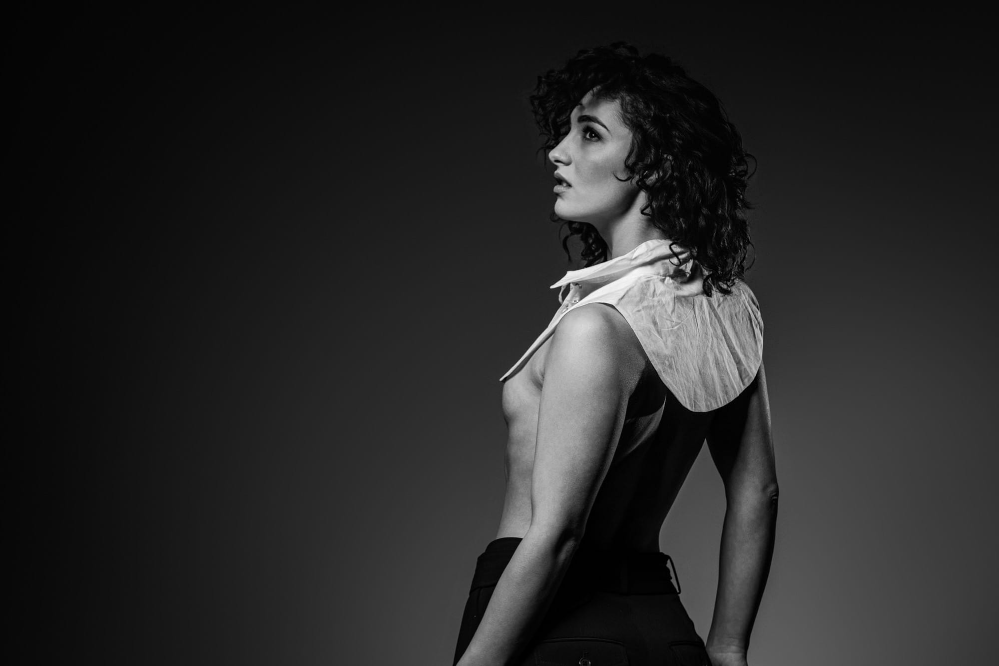 Black and white studio portrait with arms bent outward at the elbows, white lace-up collar and black trousers, nude torso, face turned to the side against a dark background.